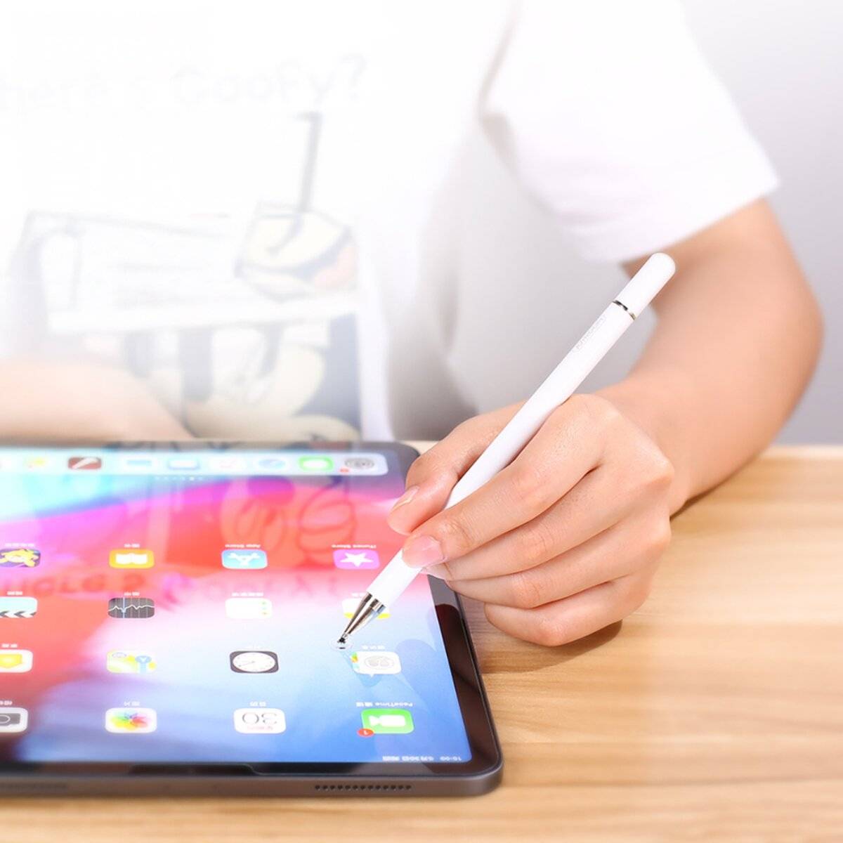 Eine Person hält einen Stylus, während sie mit einem iPad interagiert, das auf einem Holztisch liegt und dessen Home-Bildschirm mit Apps gezeigt wird.
