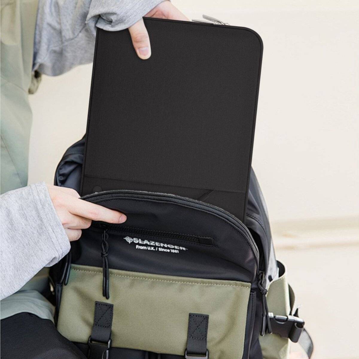 Eine Person hält ein Tablet über einem offenen Rucksack, während eine Hand auf die vordere Tasche zeigt und eine Nutzung oder Lagerung andeutet.