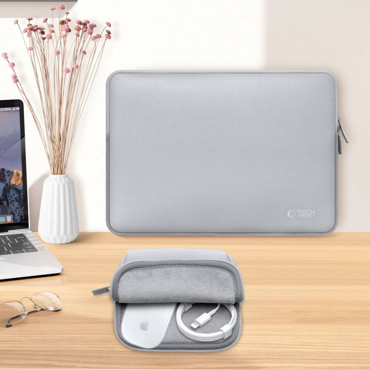 Eine silberne Laptop-Schutzhülle und ein Zubehörbeutel werden auf einer hellen Oberfläche angezeigt. Ein geschlossener Laptop, eine Brille und eine Vase mit rosa Blumen sind in der Nähe.