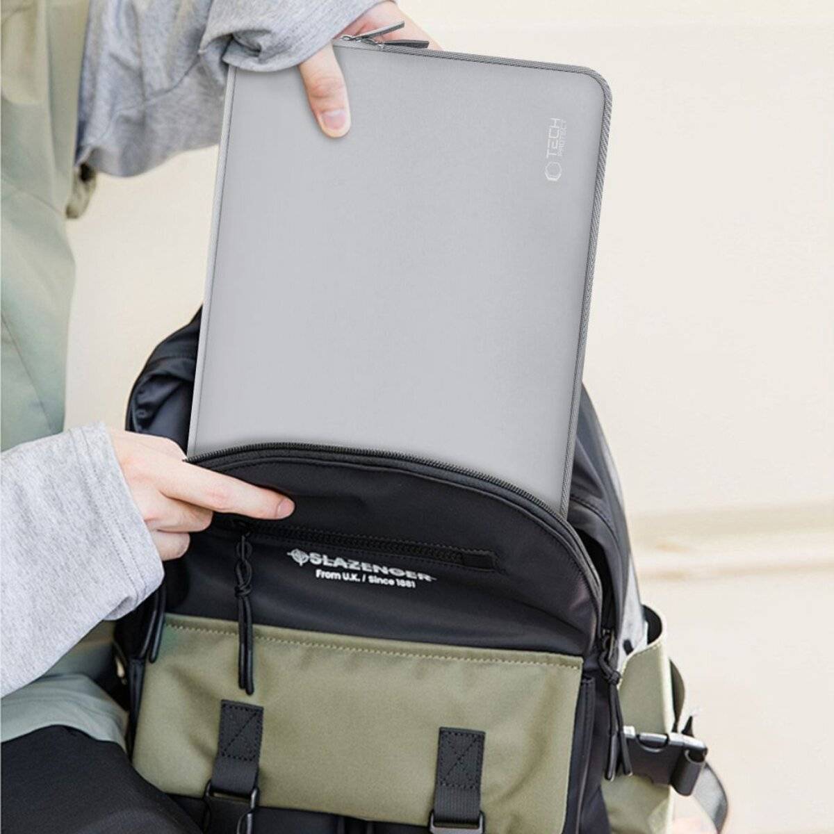 Eine Person legt ein graues Tablet in einen schwarzen und olivgrünen Rucksack. Die Person trägt einen hellgrauen Pullover und eine Jacke.