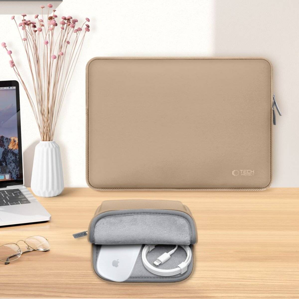 Eine beige Laptop-Tasche und ein graues Tech-Täschchen liegen auf einem Holzschreibtisch neben einem Laptop, einer Brille und einer Vase mit getrockneten Blumen. Das Täschchen ist aufgezogen und zeigt Kabel im Inneren.