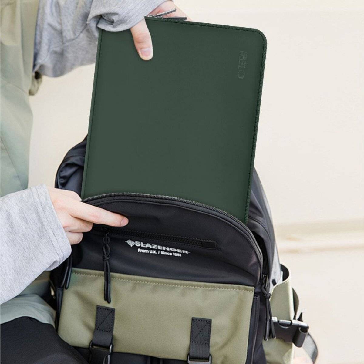 Eine Person legt ein Tablet in einen schwarzen Rucksack. Das Tablet befindet sich in einer dunkelgrünen Schutzhülle. Der Rucksack hat mehrere Taschen und Riemen.