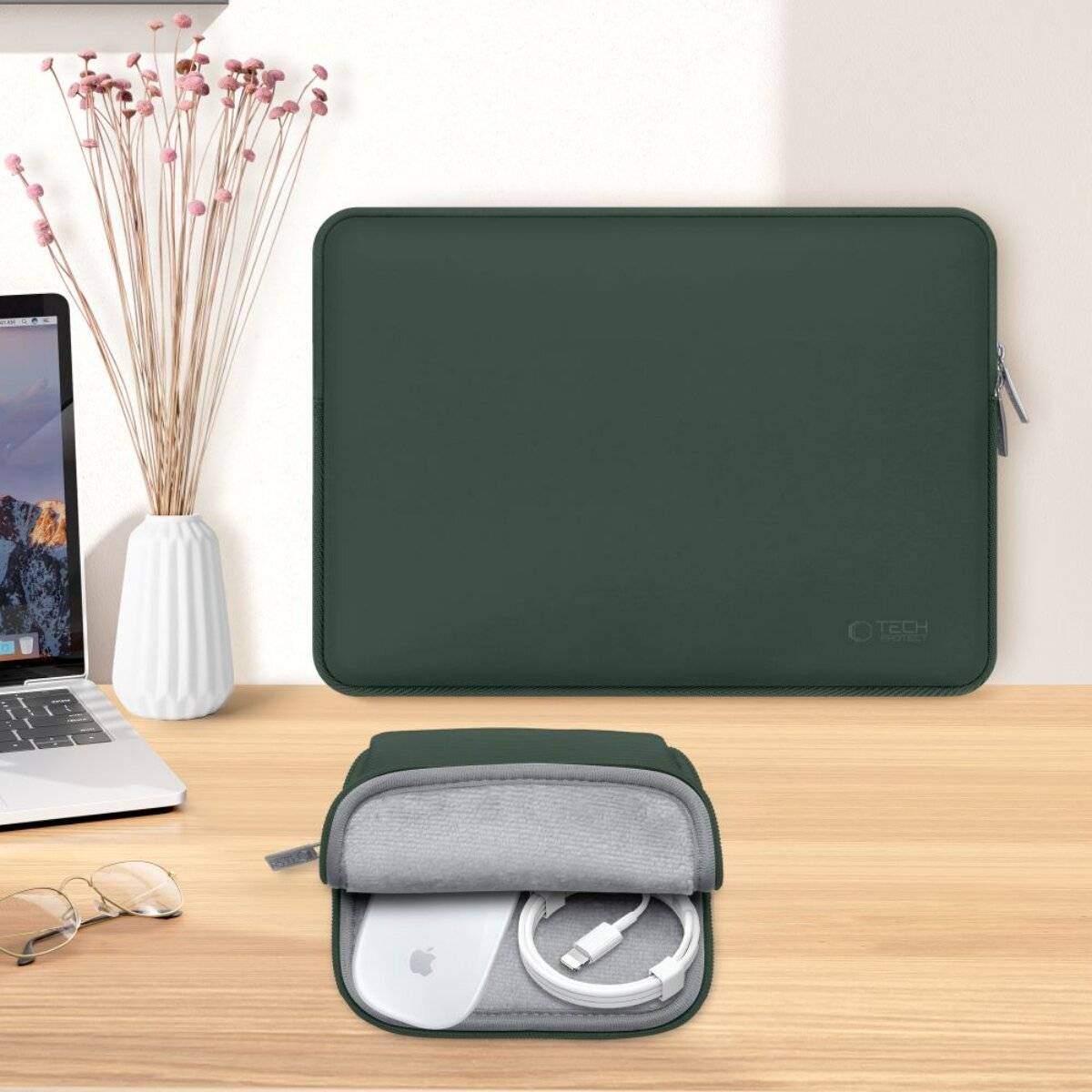 Laptop-Schutzhülle und kleine Tasche auf einem Schreibtisch; die Hülle ist grün, mit Reißverschluss versehen und zum Schutz ausgelegt. Ein Laptop, eine Brille und eine Vase sind in der Nähe.