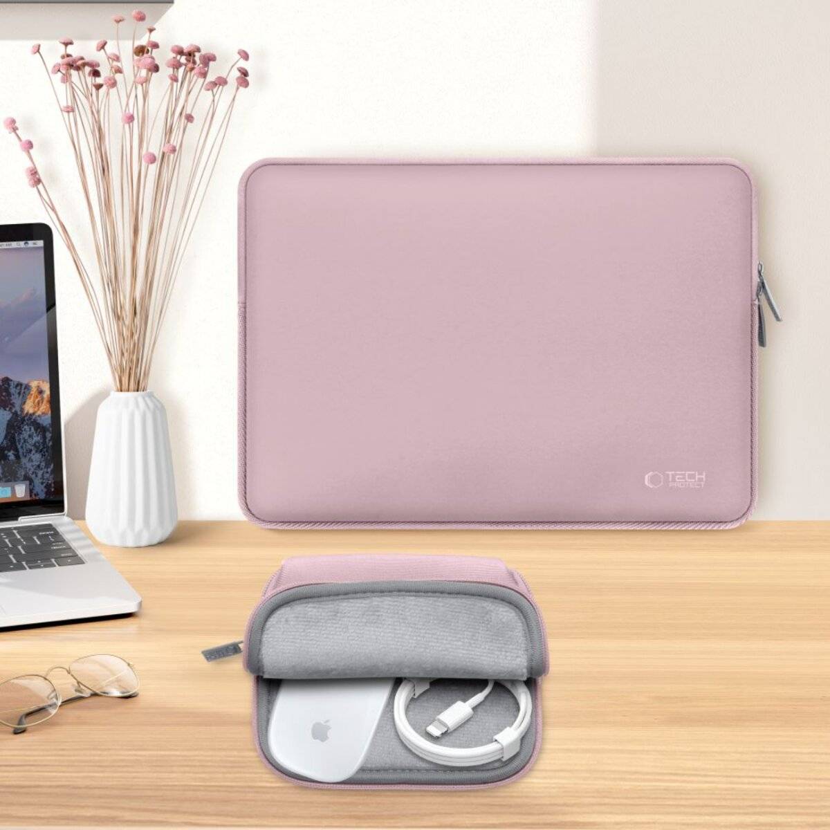 Eine hellrosa Laptoptasche auf einem Schreibtisch, neben Brille, Kabeln und einem Laptop. Eine kleine weiße Vase mit rosa Blumen ergänzt die Dekoration.