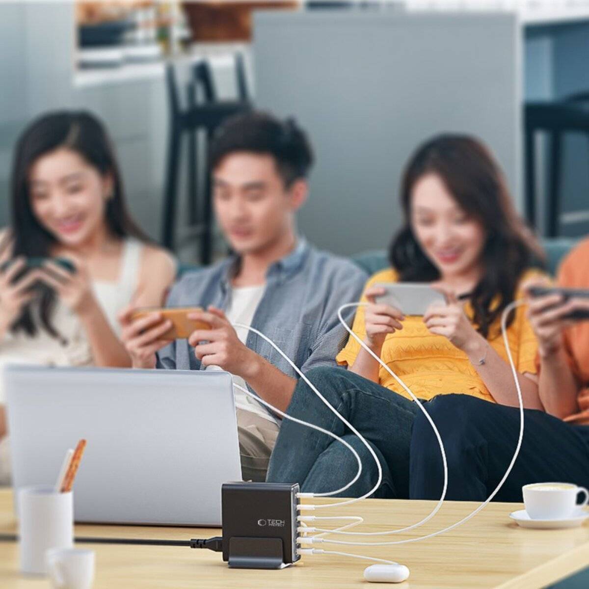 Vier Personen sitzen an einem Tisch und nutzen Smartphones, während ein Multi-Port-USB-Ladegerät ihre Geräte mit Strom versorgt. Ein Laptop und eine Kaffeetasse sind ebenfalls sichtbar.