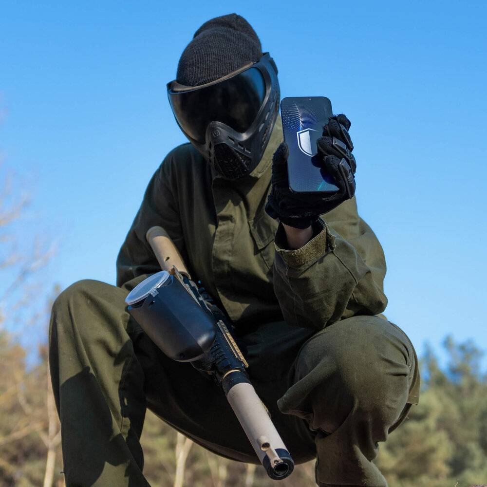 Eine Person, die eine Paintball-Maske und Ausrüstung trägt, hält ein Smartphone in einer Hand und einen Paintball-Marker in der anderen Hand, im Freien.