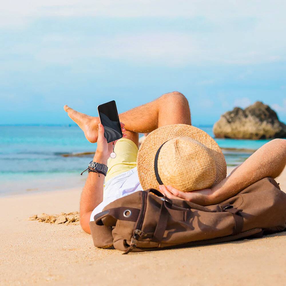 Eine Person, die auf einem sandigen Strand liegt und ein Smartphone hält. Sie trägt einen Strohhut und hat einen Rucksack neben sich, mit dem Meer im Hintergrund.