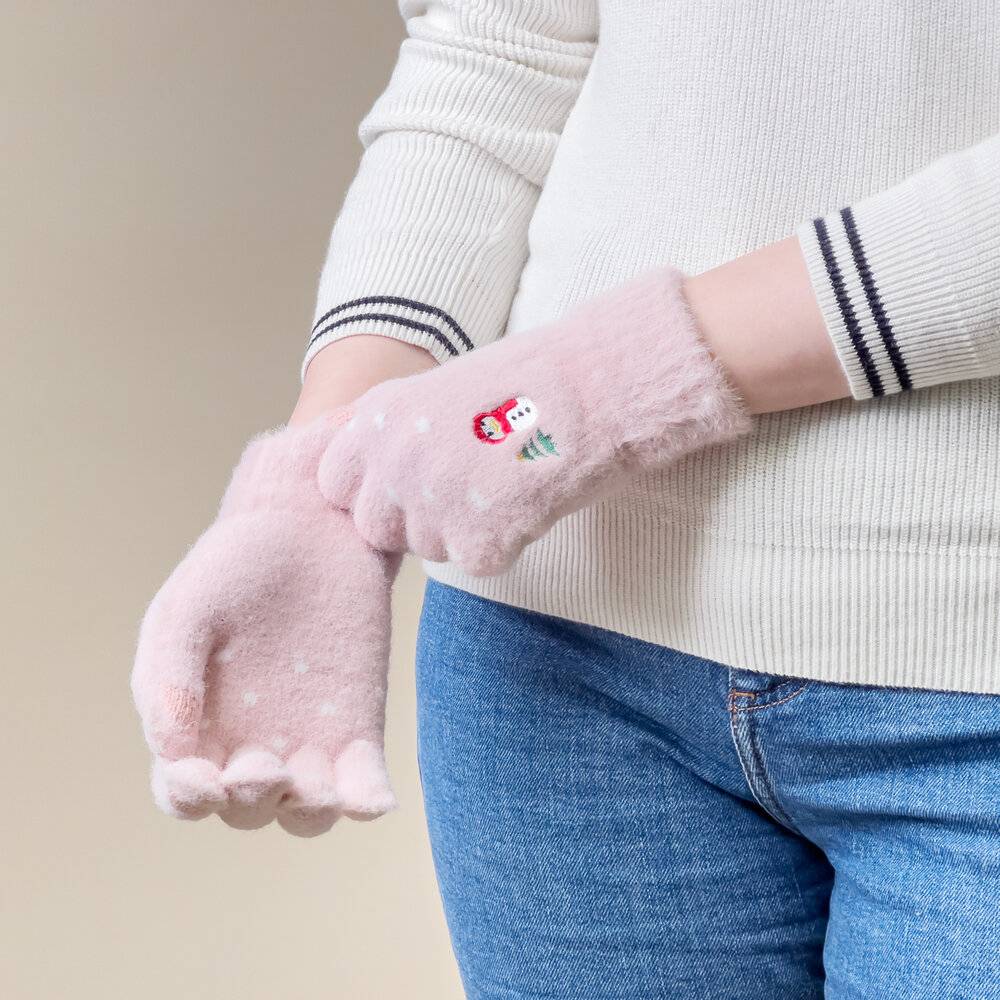 Eine Person in einem weißen Pullover und blauen Jeans hält das rechte Handgelenk mit der linken Hand, während sie rosafarbene Handschuhe mit einem kleinen roten bestickten Design trägt.