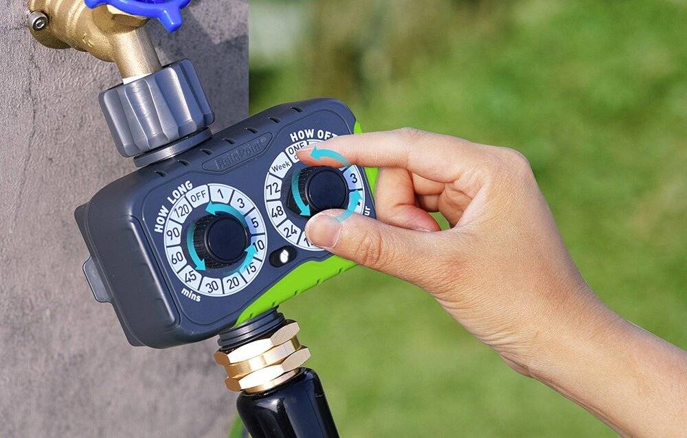 Eine Hand stellt einen Bewässerungstimer mit zwei Wählscheiben ein, der an einem Gartenhahn befestigt ist, mit Einstellungen für „Wie lange