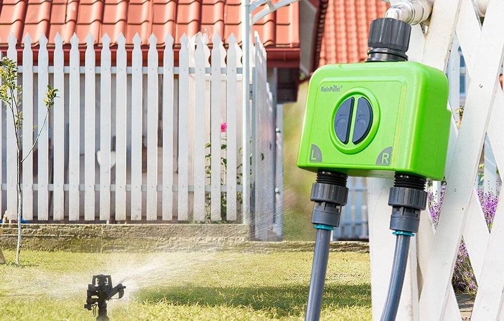 Ein Sprinkler bewässert einen Rasen vor einem weißen Lattenzaun. Ein grüner Bewässerungstimer ist am Schlauch befestigt und so eingestellt, dass er das Gebiet bewässert.