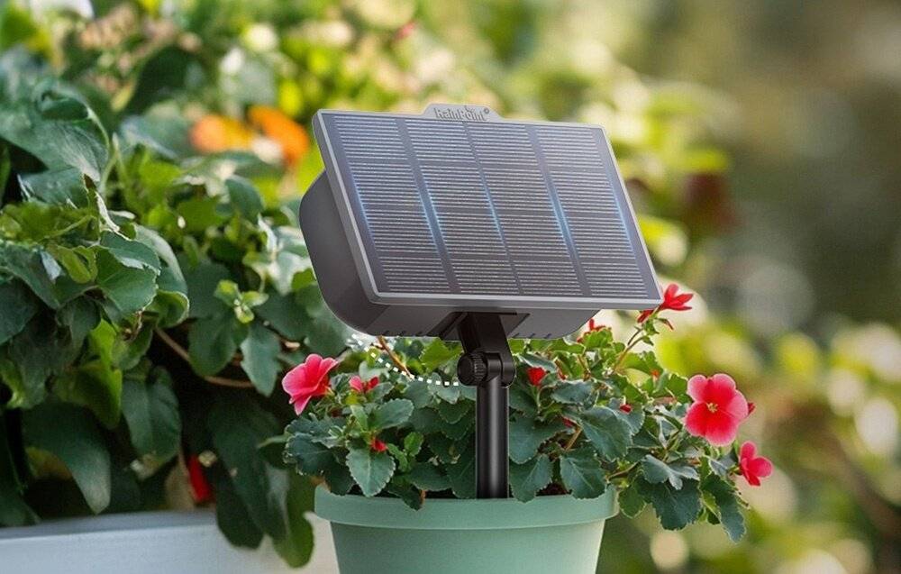 Ein kleines Solarpanel auf einem Pfahl montiert wird in einem Blumentopf platziert, der von bunten Blumen umgeben ist, was auf eine Gartenumgebung hindeutet.