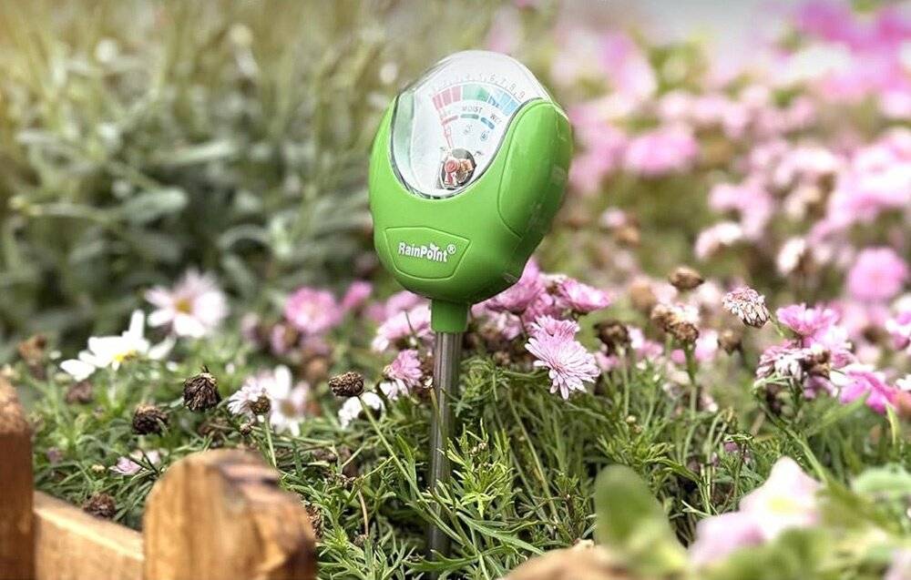 Ein Bodenfeuchtemessgerät mit grünem Gehäuse wird in die Erde zwischen blühenden rosa Blumen eingeführt und misst die Bodenfeuchtewerte.