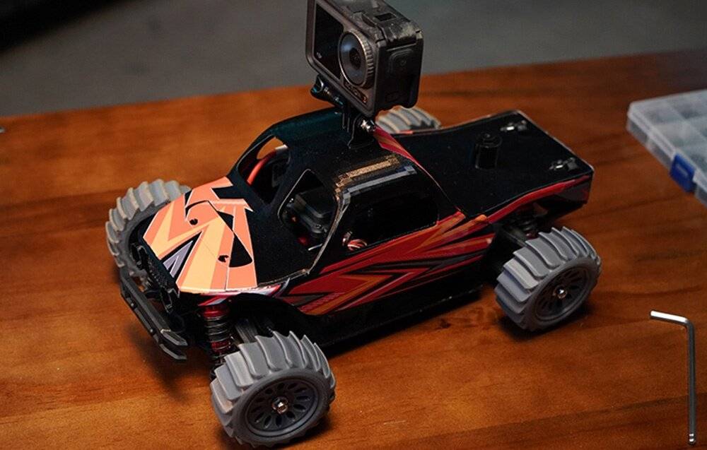 Ein ferngesteuertes Auto mit großen Reifen und einem schwarz-orangefarbigen Design hat eine auf der Oberseite montierte Kamera, die auf einer Holzoberfläche zu sehen ist.
