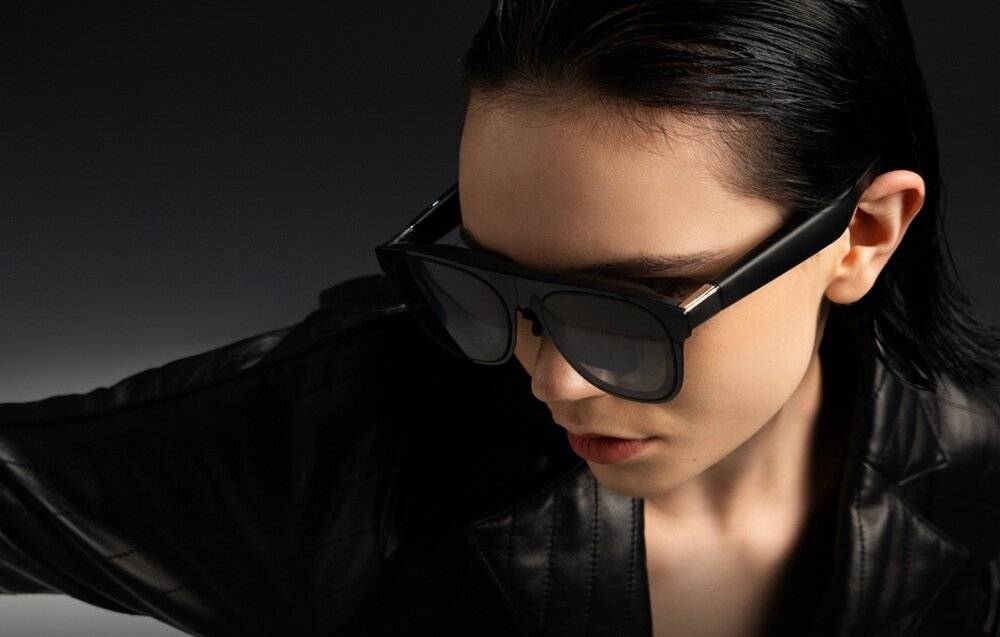 Eine Person mit Sonnenbrille und Lederjacke erscheint in einer schattierten Umgebung und blickt nachdenklich nach unten.