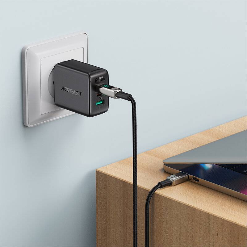 Ein schwarzes USB-Ladegerät ist in eine Wandsteckdose eingesteckt, mit einem Kabel, das mit einem Laptop auf einem Holzschreibtisch verbunden ist.