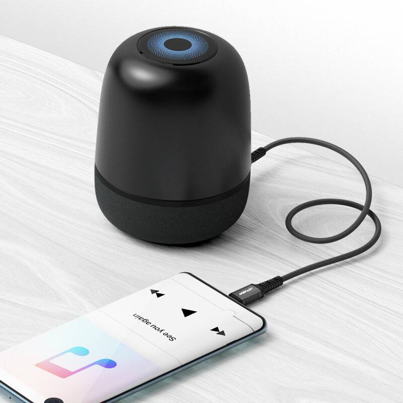 Ein schwarzer tragbarer Lautsprecher, der mit einem Smartphone verbunden ist, das eine Musik-App auf einer Holzoberfläche anzeigt.