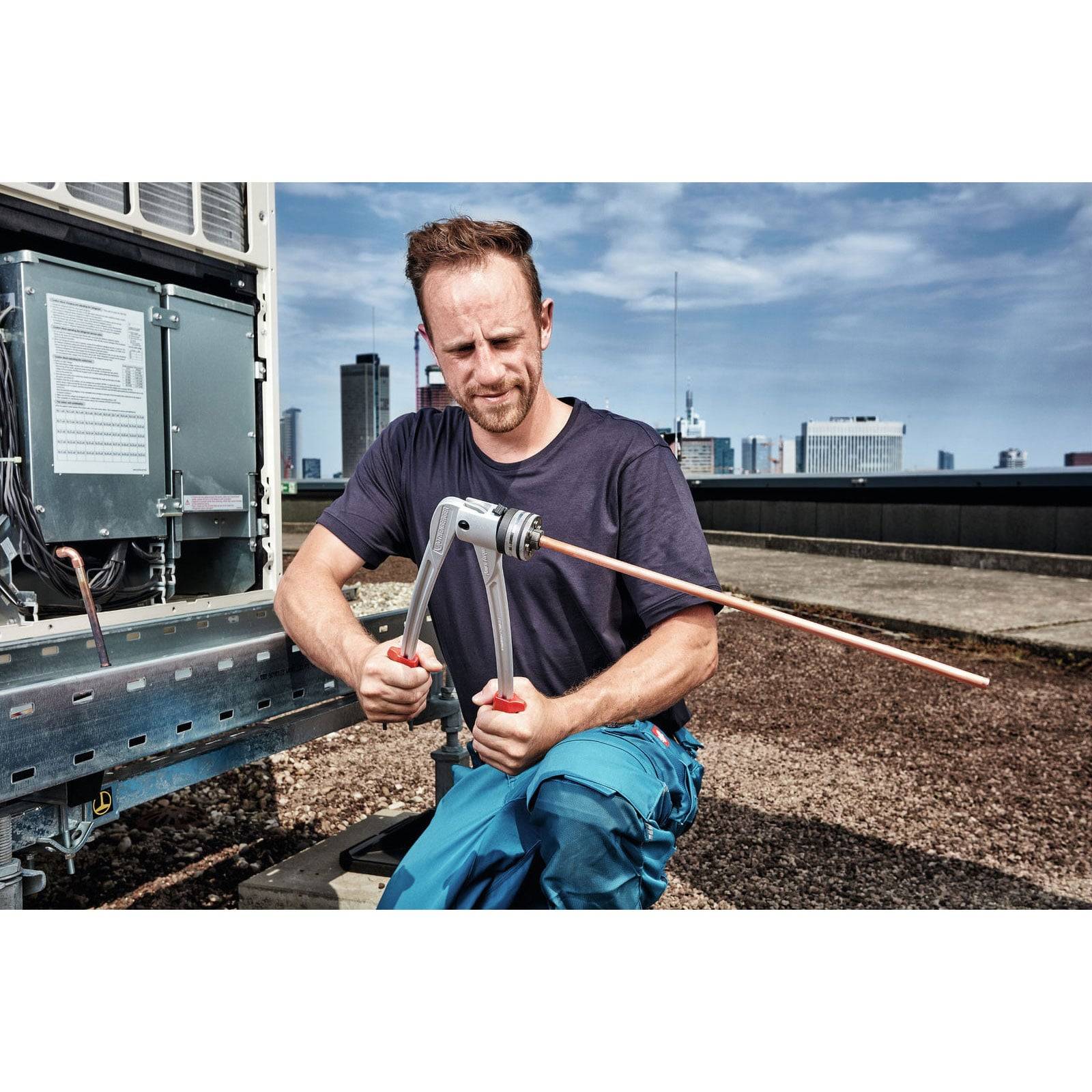 Ein Techniker in blauen Hosen benutzt ein Werkzeug auf einer Dachklima-Einheit, während im Hintergrund Stadtgebäude zu sehen sind, und führt Wartungsarbeiten durch.