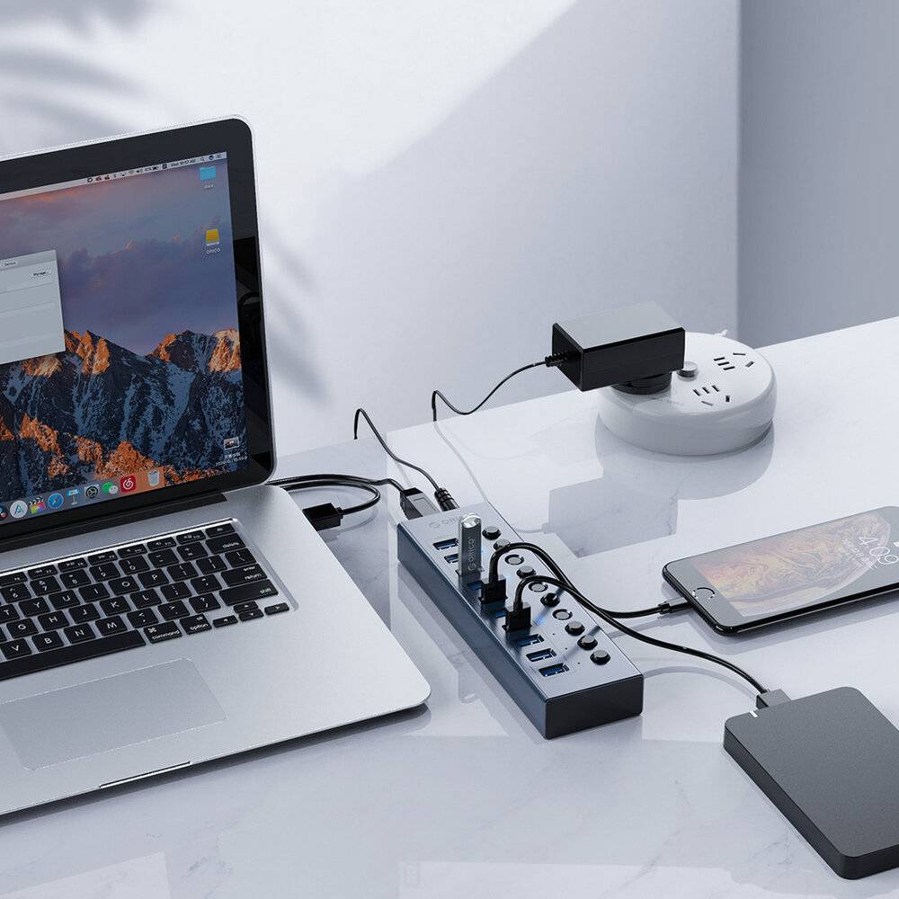 Ein Laptop, der an eine Steckdosenleiste angeschlossen ist, mit einem sich in der Nähe aufladenden Smartphone. Das Setup deutet auf einen Arbeitsplatz oder eine Ladestation hin.