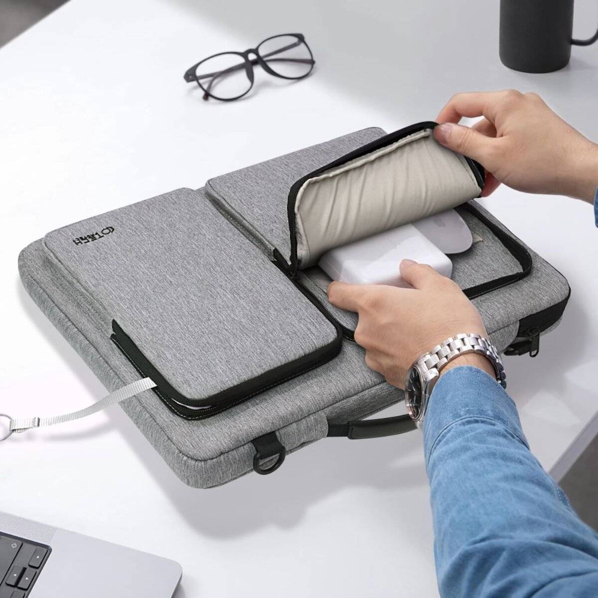 Eine Person legt ein Smartphone in eine graue Laptoptasche mit organisierten Fächern auf einem Tisch. Eine Brille und eine Tasse sind in der Nähe.