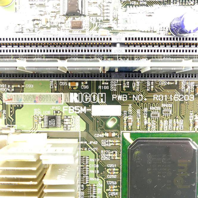 Nahaufnahme einer Computermainboard mit einem Ricoh-Chip, Anschlüssen und einem Kühlkörper, die Schaltungspuren und elektronische Komponenten zeigt.