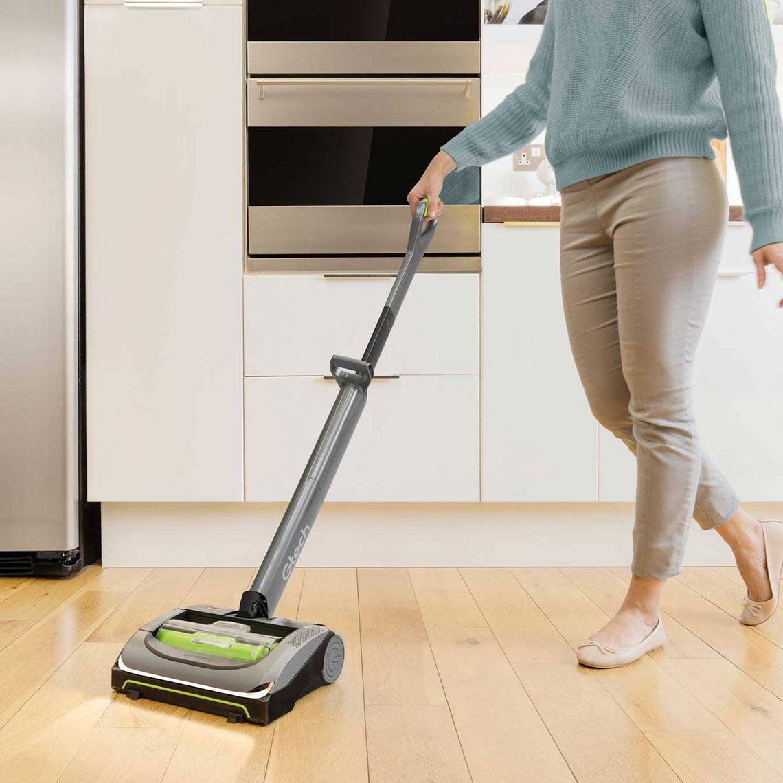 Eine Person saugt einen Holzboden mit einem kabellosen Staubsauger in einer modernen Küche mit hellkabinetten und Edelstahlgeräten ab.