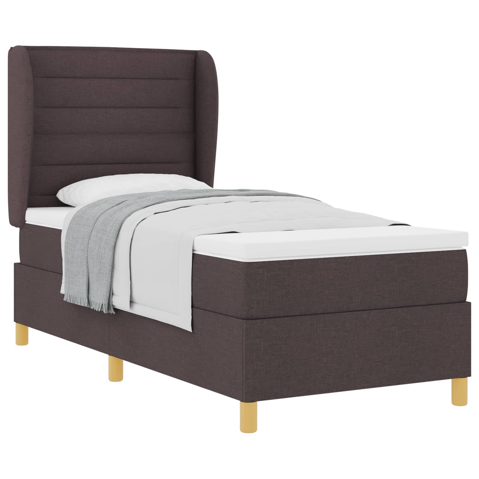 Ein Einzelbett mit dunklem Stoff-Kopfteil, weißer Matratze und einer grau gefalteten Decke ordentlich auf der Oberseite. Das Bett hat Holzbeine.