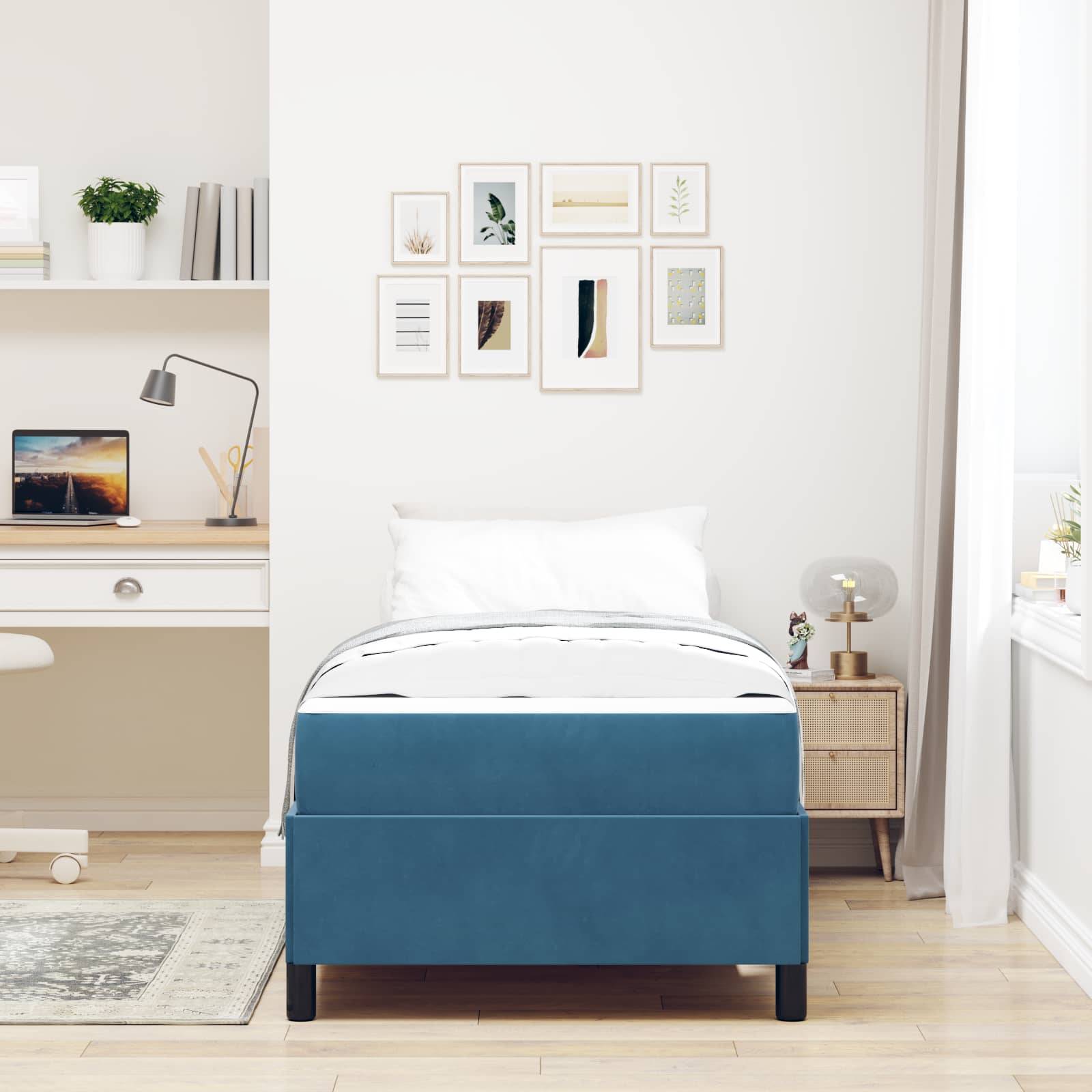 Ein minimalistisches Schlafzimmer und Arbeitsbereich mit einem blauen Bett, einem Nachttisch, einem Schreibtisch mit offenem Laptop, einem Bücherregal und Wandkunst.