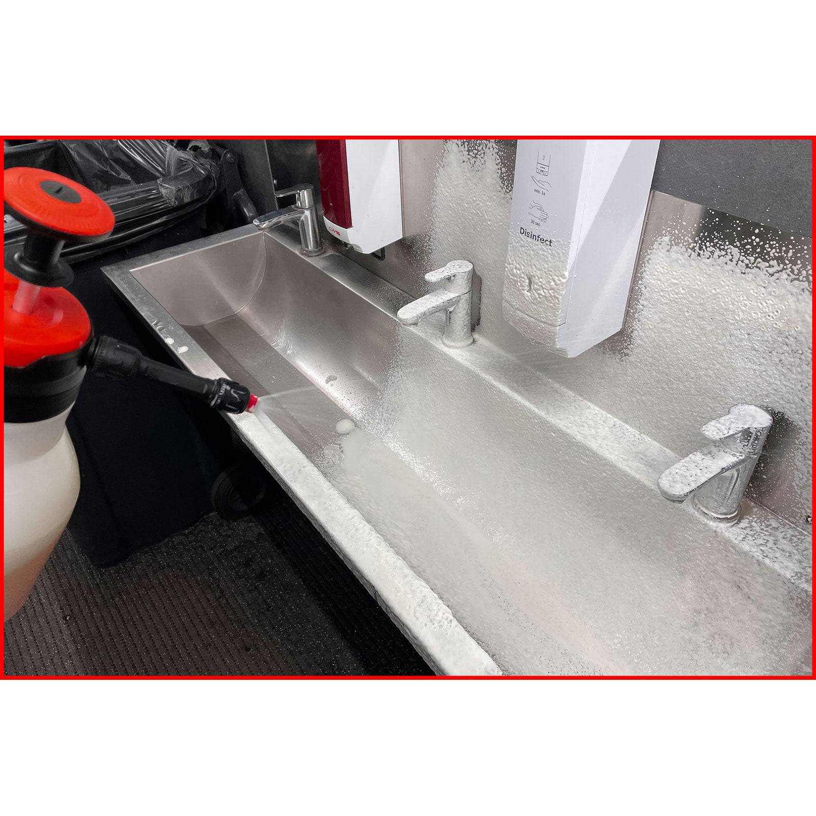 Ein langer Edelstahlspülbecken, der mit Reinigungslösung aus einer rot-weißen Sprühflasche besprüht wird, mit zwei Seifenspendern oben.