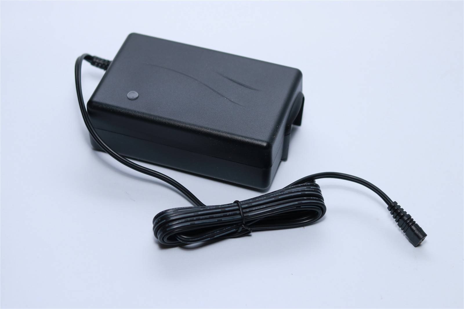 Ein schwarzes Netzteil mit einem langen, spiralförmigen Kabel liegt auf einer flachen Oberfläche und verfügt über eine kleine, kreisförmige Kontrollleuchte auf der Oberseite.