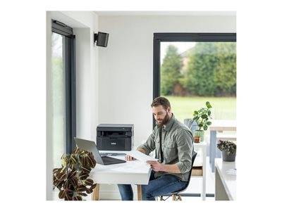 Ein Mann sitzt an einem weißen Schreibtisch mit einem Laptop und hält ein Blatt Papier in der Hand. Ein Drucker und eine Pflanze befinden sich in der Nähe; große Fenster bieten einen Blick nach draußen.
