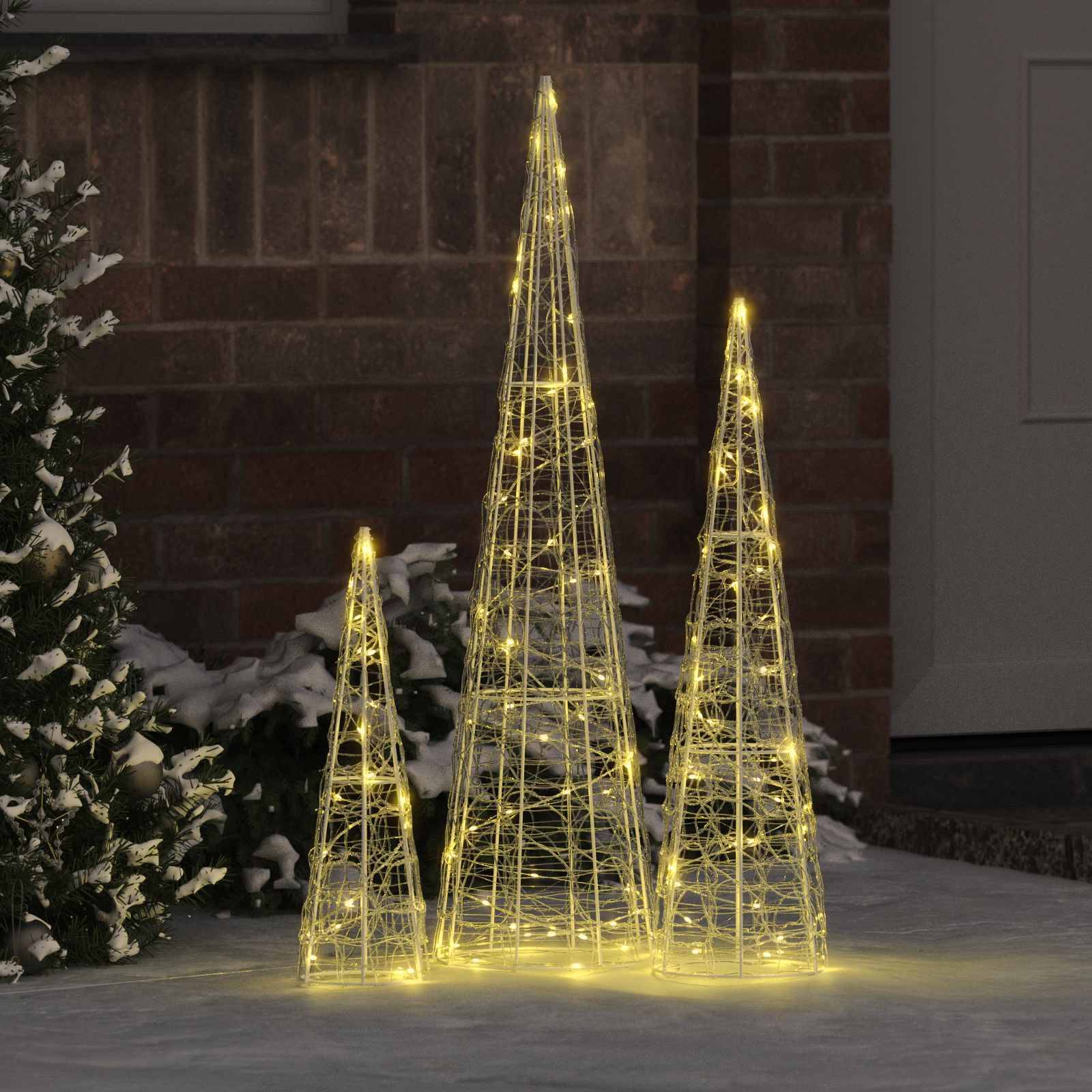 Drei beleuchtete Drahtgitter-Kegelbäume, die mit warmweißen Lichtern geschmückt sind, sind auf einer verschneiten Außenfläche in der Nähe eines kleinen Immergrünstrauchs angeordnet.