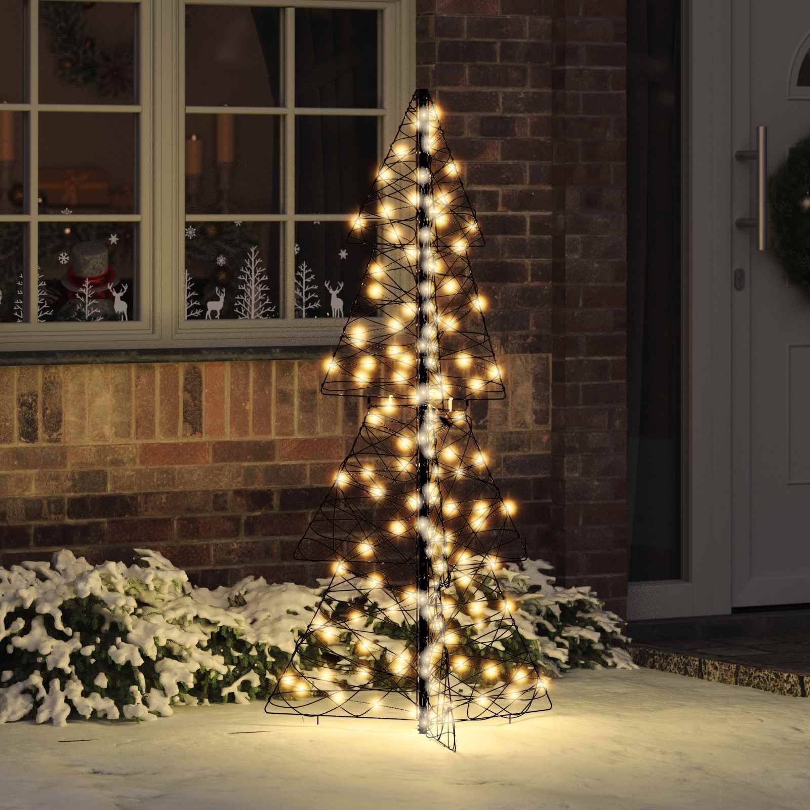Ein dekorativer Drahtgitter-Weihnachtsbaum mit leuchtenden Lichtern steht vor einem Haus, umgeben von Schnee, mit Feiertagsaufklebern am Fenster.