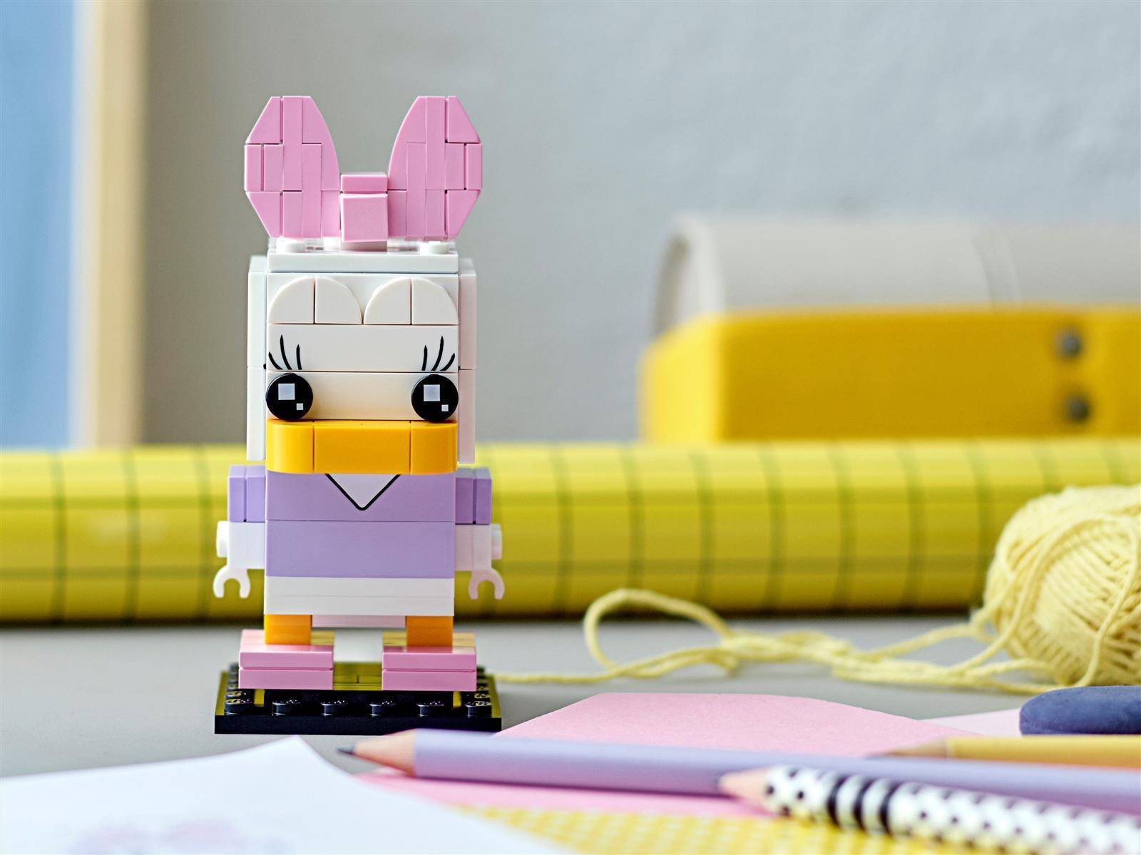 Eine kleine LEGO-ähnliche Hasenfigur mit rosa Ohren und violettem Outfit steht auf einem Tisch, mit Bastelvorräten und gerolltem Papier im Hintergrund.