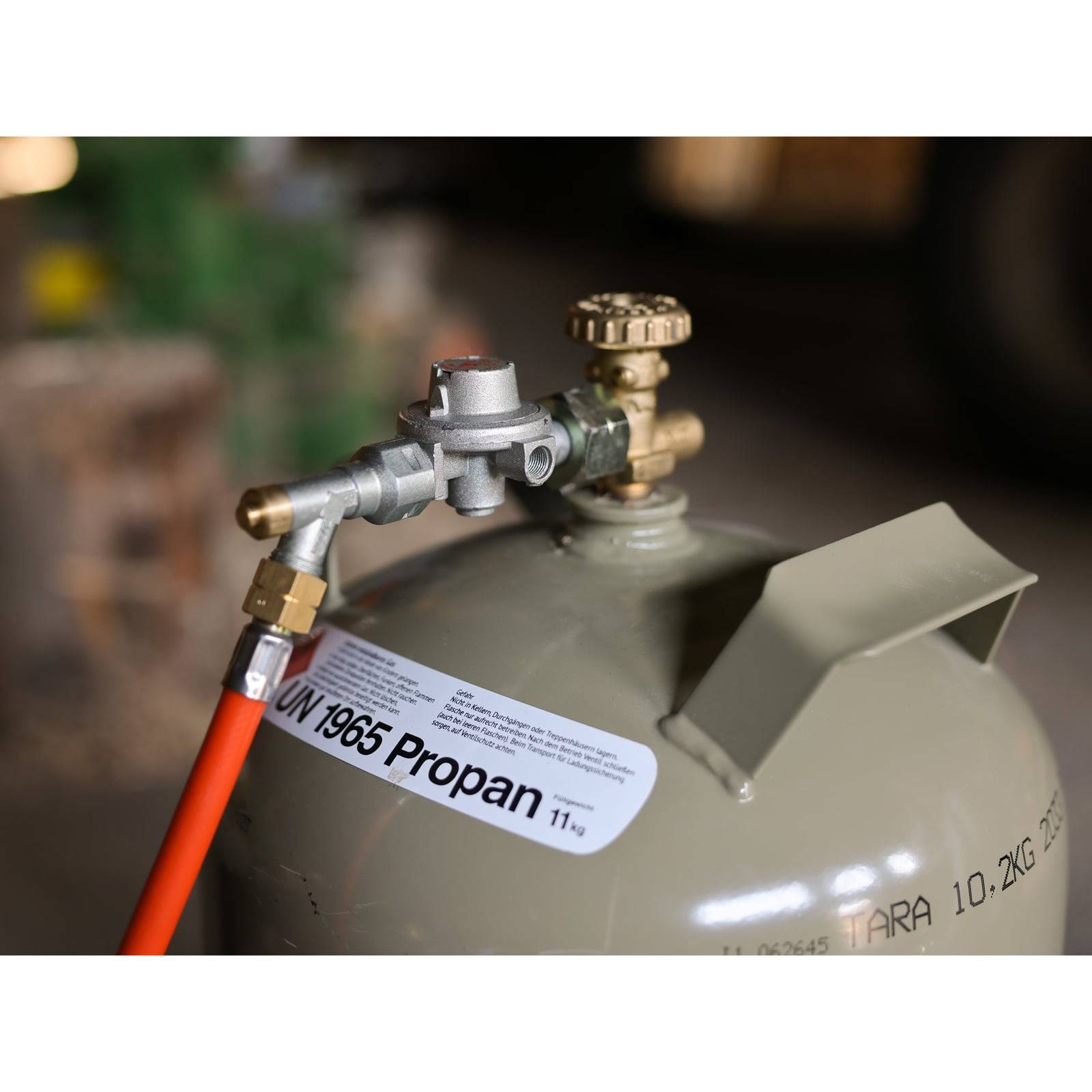 Eine Propangasflasche mit Druckregler und rotem Schlauch, gekennzeichnet mit 'UN 1965 Propan 11kg'.
