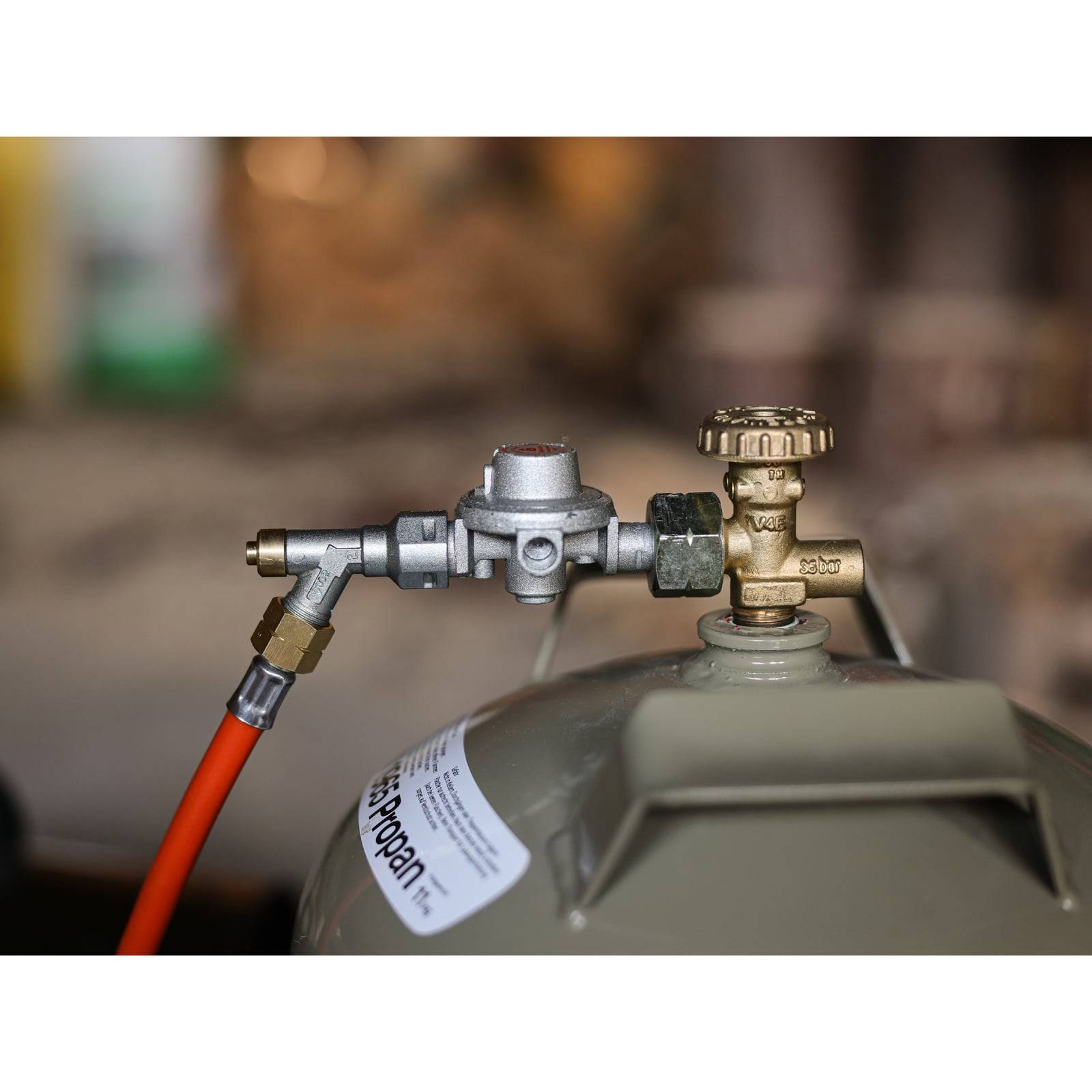 Eine Propangasflasche mit angeschlossenem orangefarbenem Schlauch und Ventil, aufgestellt in einer Werkstattumgebung. Das Etikett der Flasche zeigt Sicherheitsinformationen.