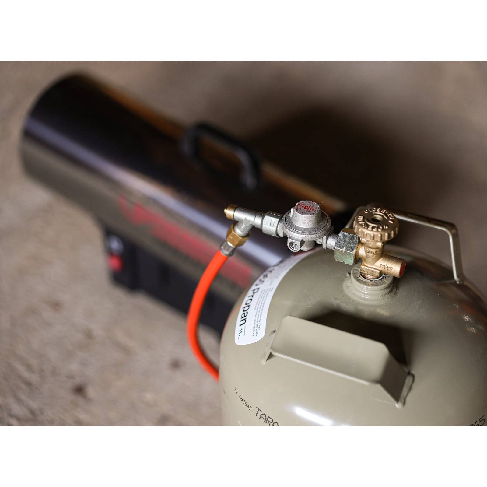 Ein Propanheizgerät, das an einen Tank angeschlossen ist, mit einem roten Ventil und einem orangefarbenen Schlauch. Der Heizer ist metallisch und zylindrisch und wahrscheinlich für den Außen- oder industriellen Einsatz konzipiert.