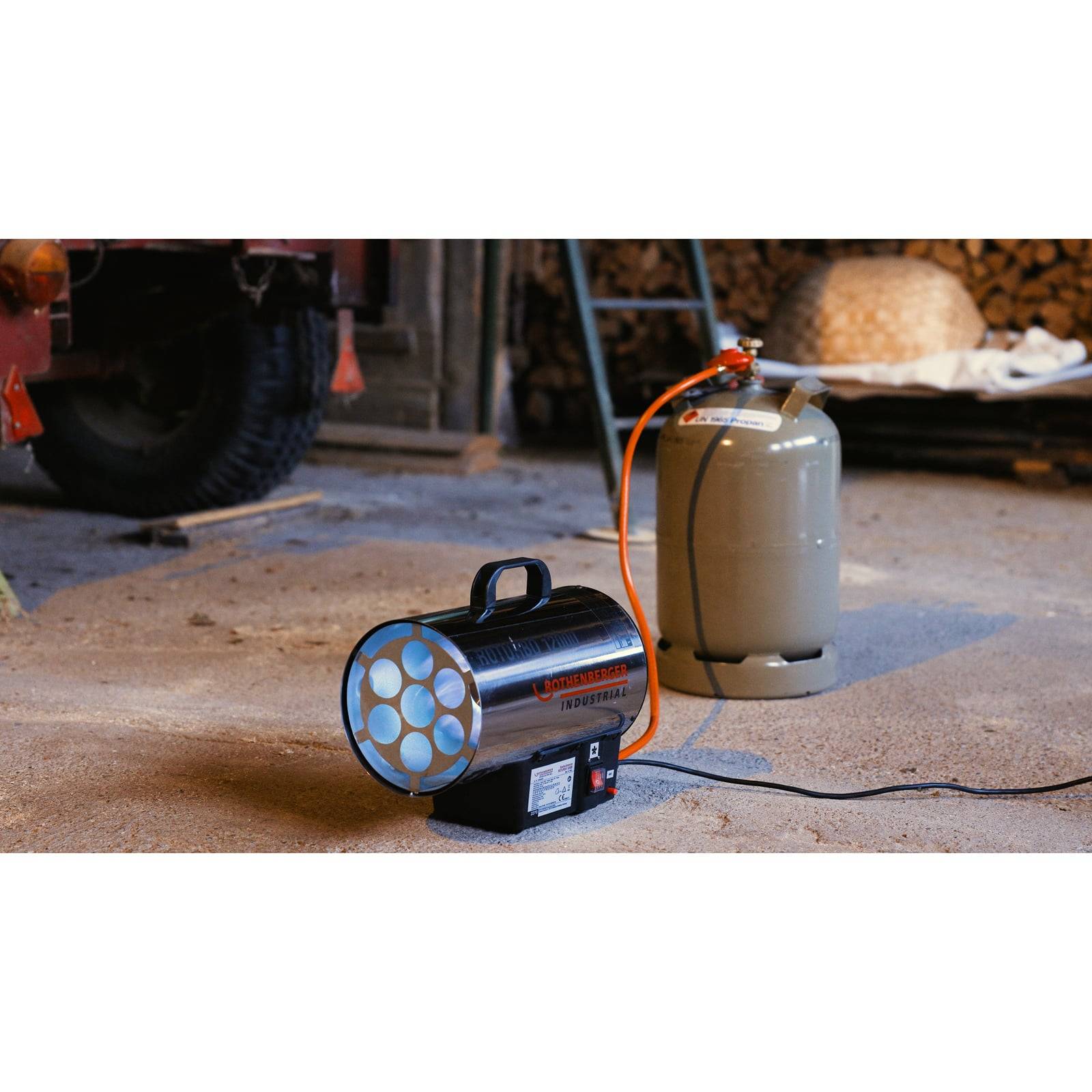 Ein tragbarer Industrieofen steht auf dem Boden und ist über einen orangefarbenen Schlauch mit einer Propangasflasche in einer Werkstattumgebung verbunden.