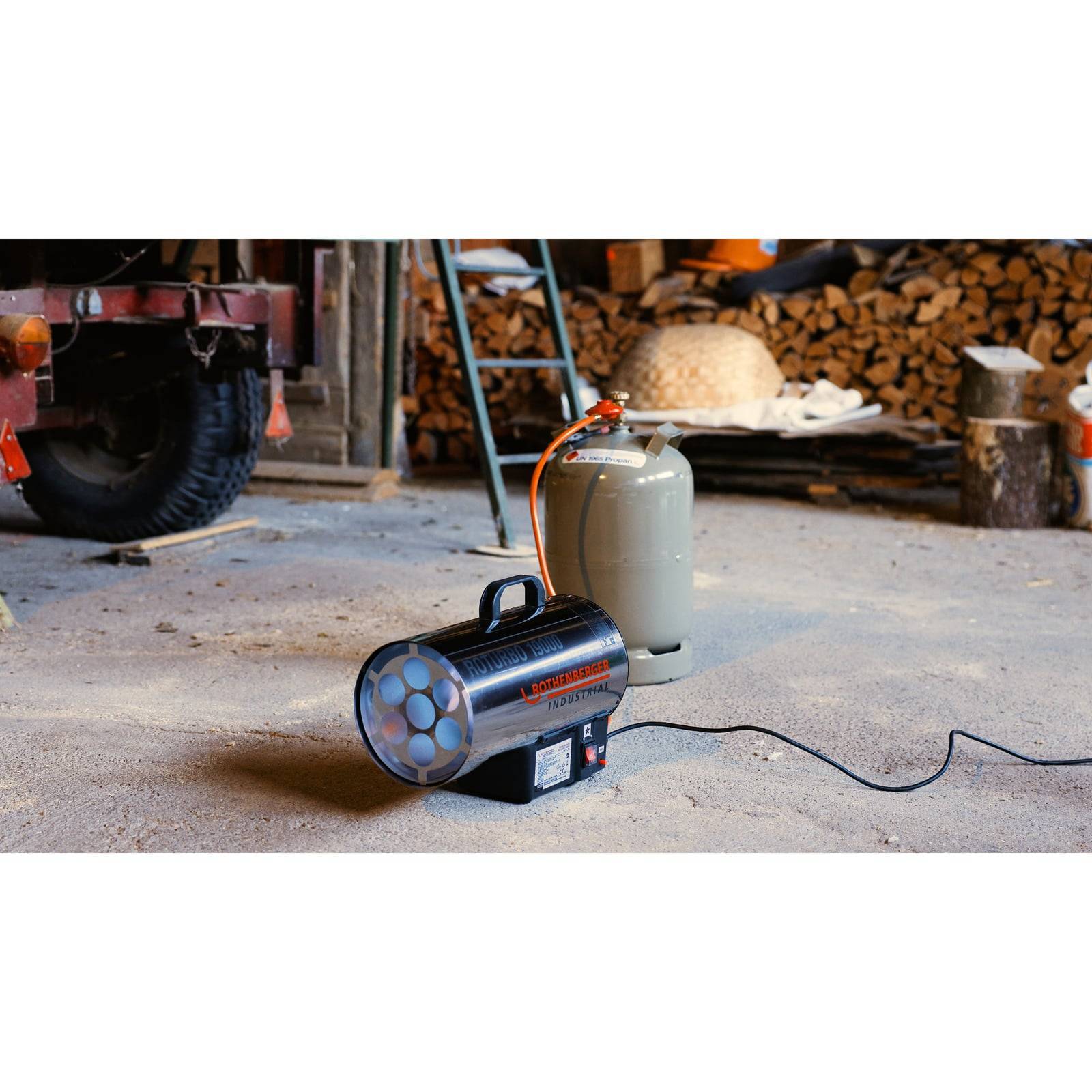 Tragbarer Gasheizöfen in einer Werkstatt, angeschlossen an einen Propantank, mit einer Leiter und gestapeltem Brennholz im Hintergrund.