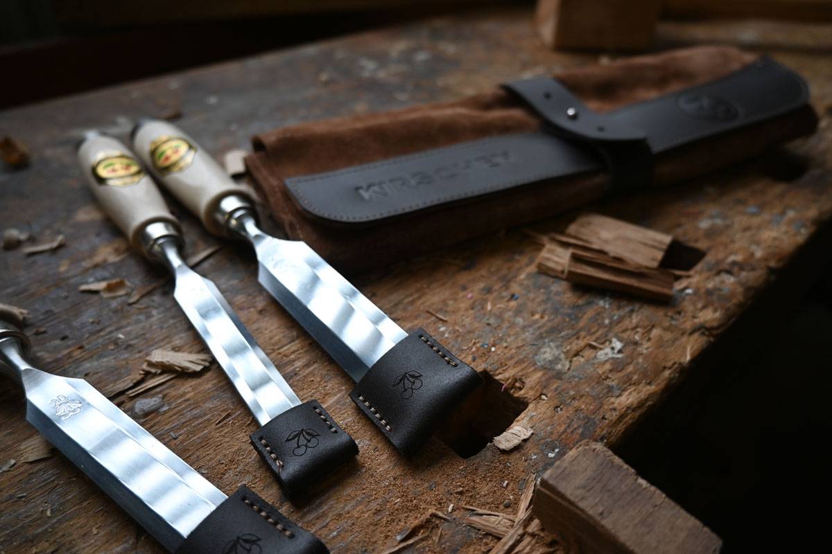 Zwei Holzbearbeitungsmeiβel mit Ledergriffen ruhen auf einem Holztisch, neben einer braunen Leder-Werkzeugrolle. Holzspäne umgeben die Werkzeuge.