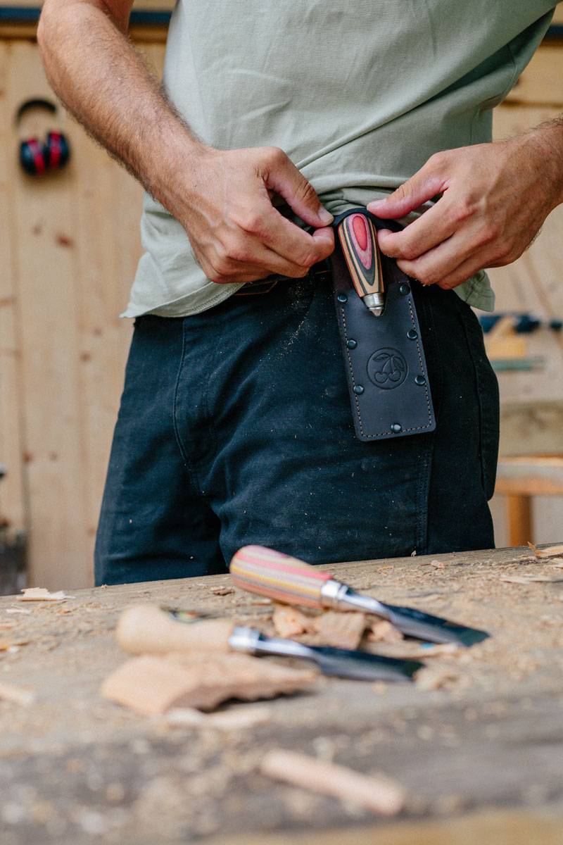 Eine Person hält ein Holzbearbeitungswerkzeug mit einer Holzklinge, das bereit ist, in eine an ihrem Gürtel befestigte Lederscheide eingeführt zu werden.