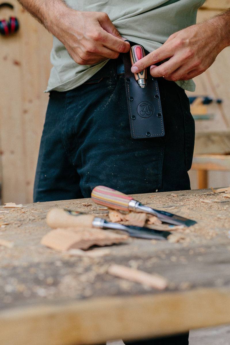 Eine Person benutzt einen Holzbearbeitungsmeisel an einem Stück Holz. Holzspäne und zusätzliche Meisel sind auf der Werkbank verstreut.