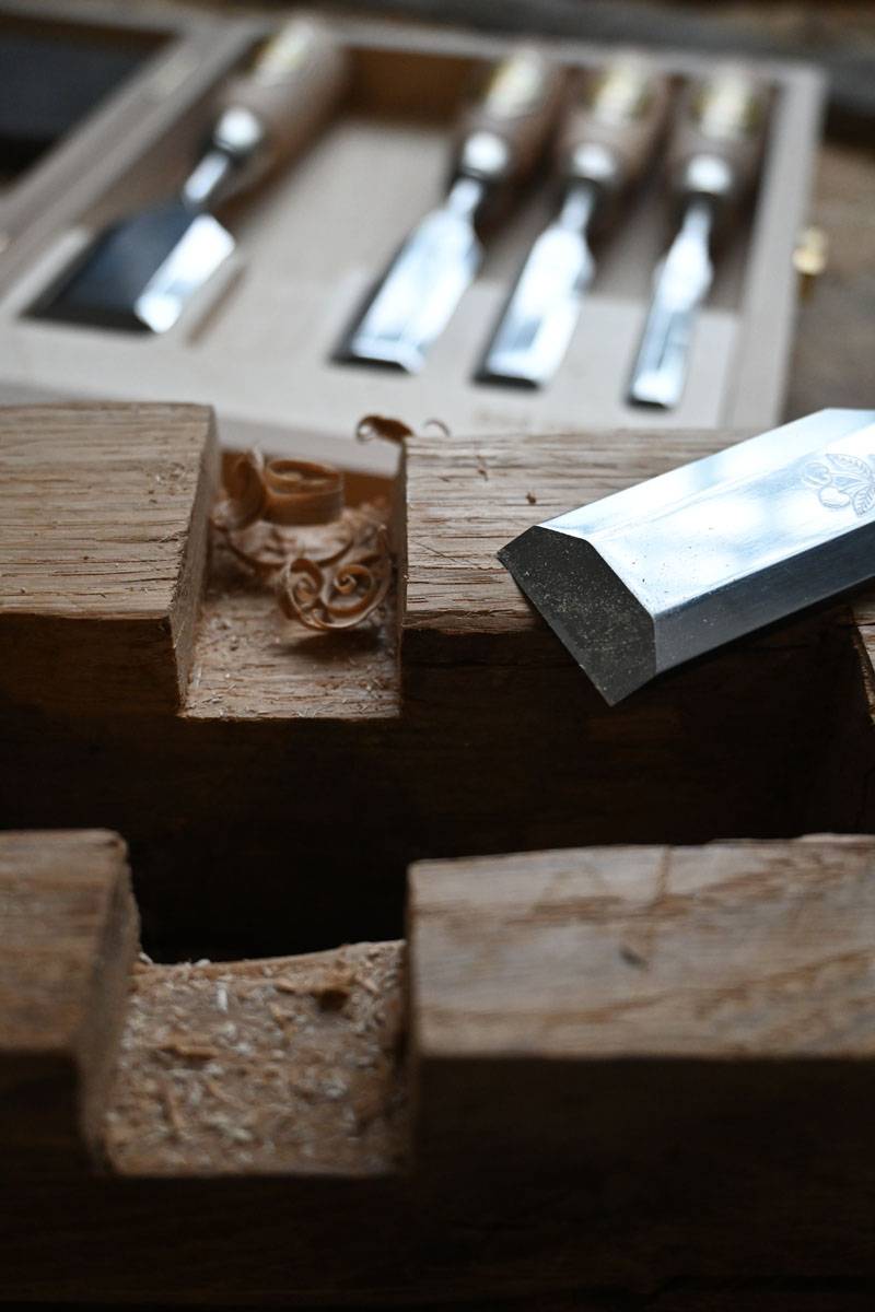 Meißel, der auf einer Holzwerkbank ruht, umgeben von Holzspänen, mit einem Satz Meißel im Hintergrund.