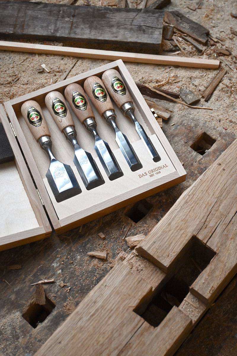 Ein Satz von sechs Holzmeißeln in einer Holzkiste auf einer Werkbank. Die Meißel haben Holzgriffe mit Etiketten und glänzende Metallklingen.