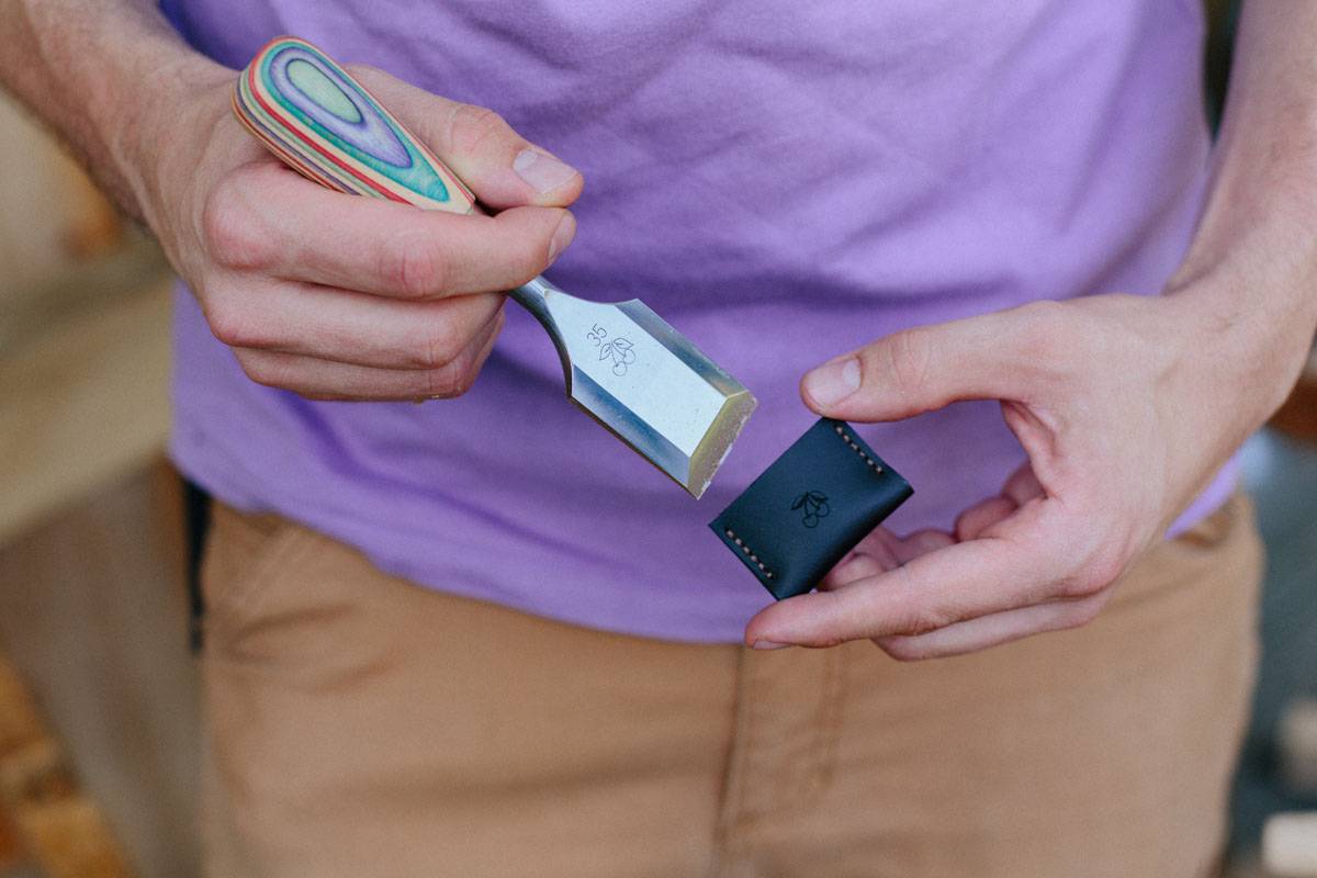 Eine Person hält ein Werkzeug mit farbigem Griff in einer Hand und ein schwarzes, rechteckiges Objekt mit kleinen Löchern in der anderen Hand, vor einem lila Hemd.