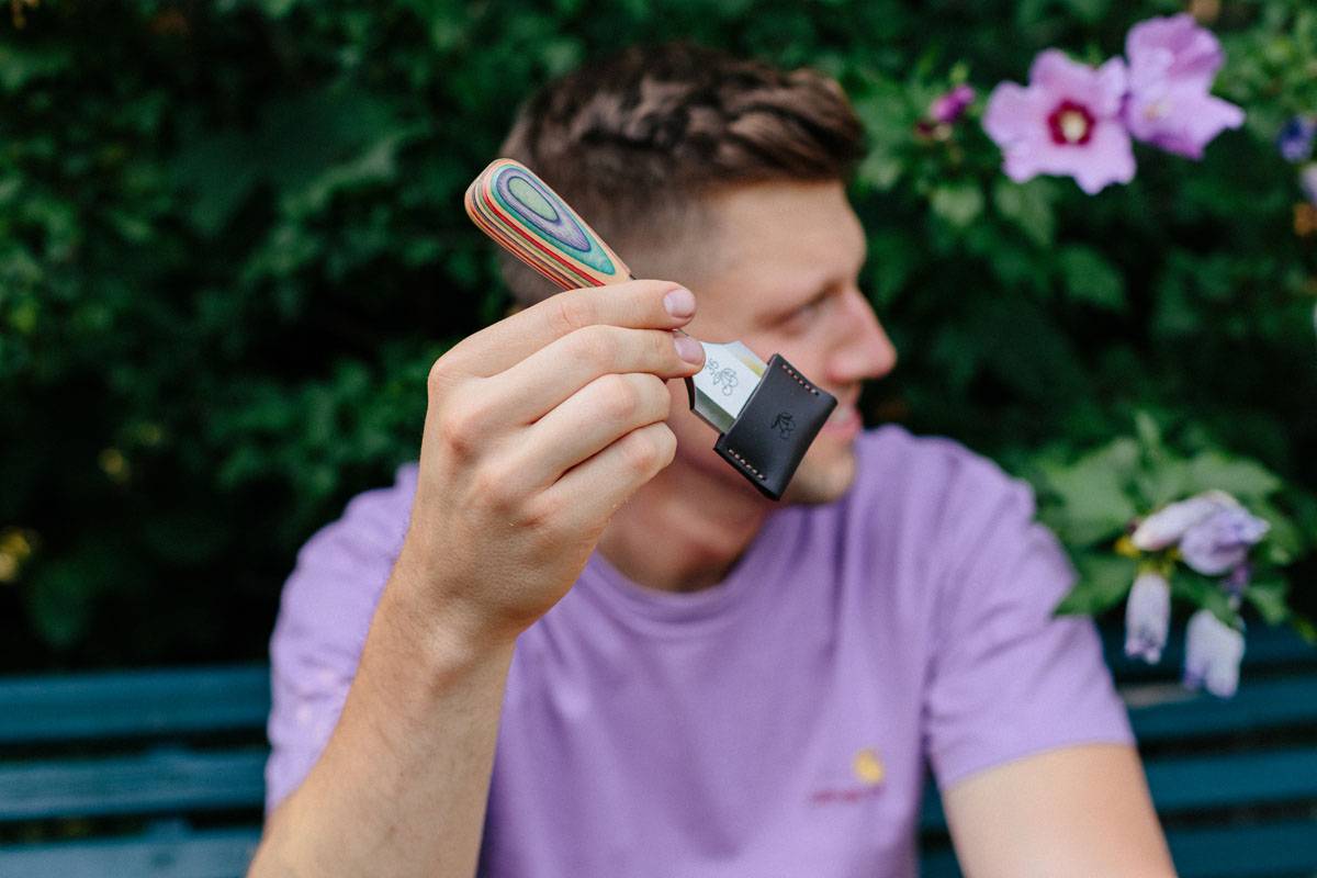 Eine Person in einem violetten Hemd hält ein kleines Campingmesser mit buntem Griff und Scheide. Grünes Laub und violette Blüten im Hintergrund.