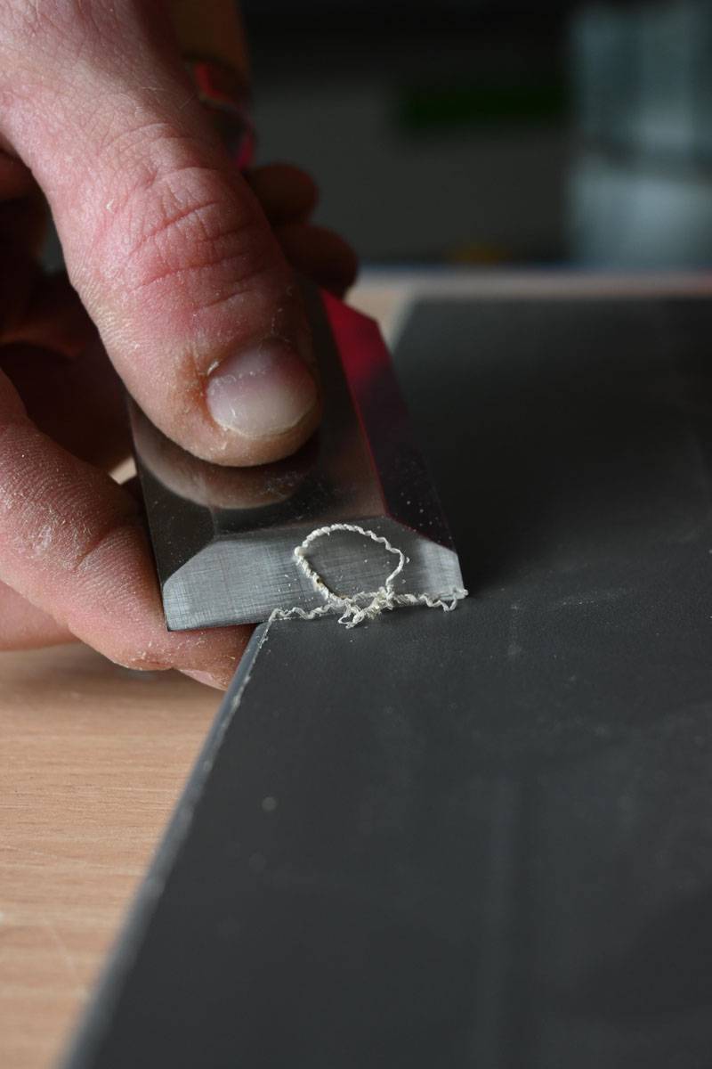 Eine Nahaufnahme einer Hand, die einen Metallschaber verwendet, um eine kleine Materialwelle von einer ebenen Oberfläche abzuschaben, möglicherweise zur Verfeinerung oder Reinigung.