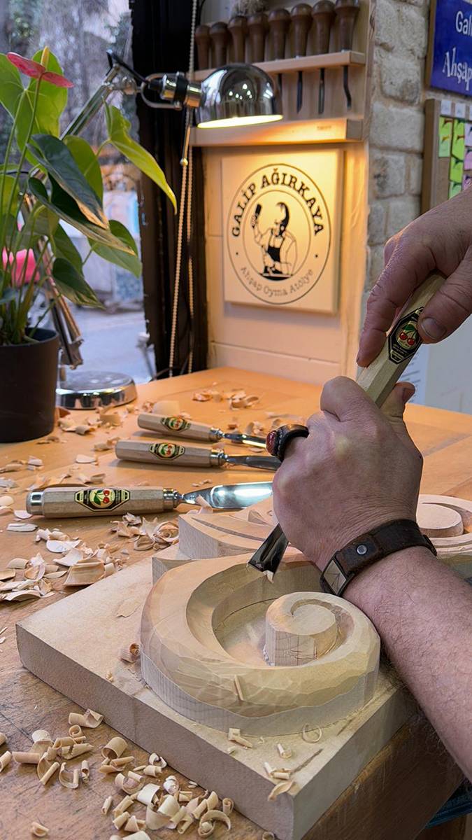 Eine Person, die mit Werkzeugen ein Holzobjekt bearbeitet, umgeben von Holzspänen in einer Werkstattsituation.