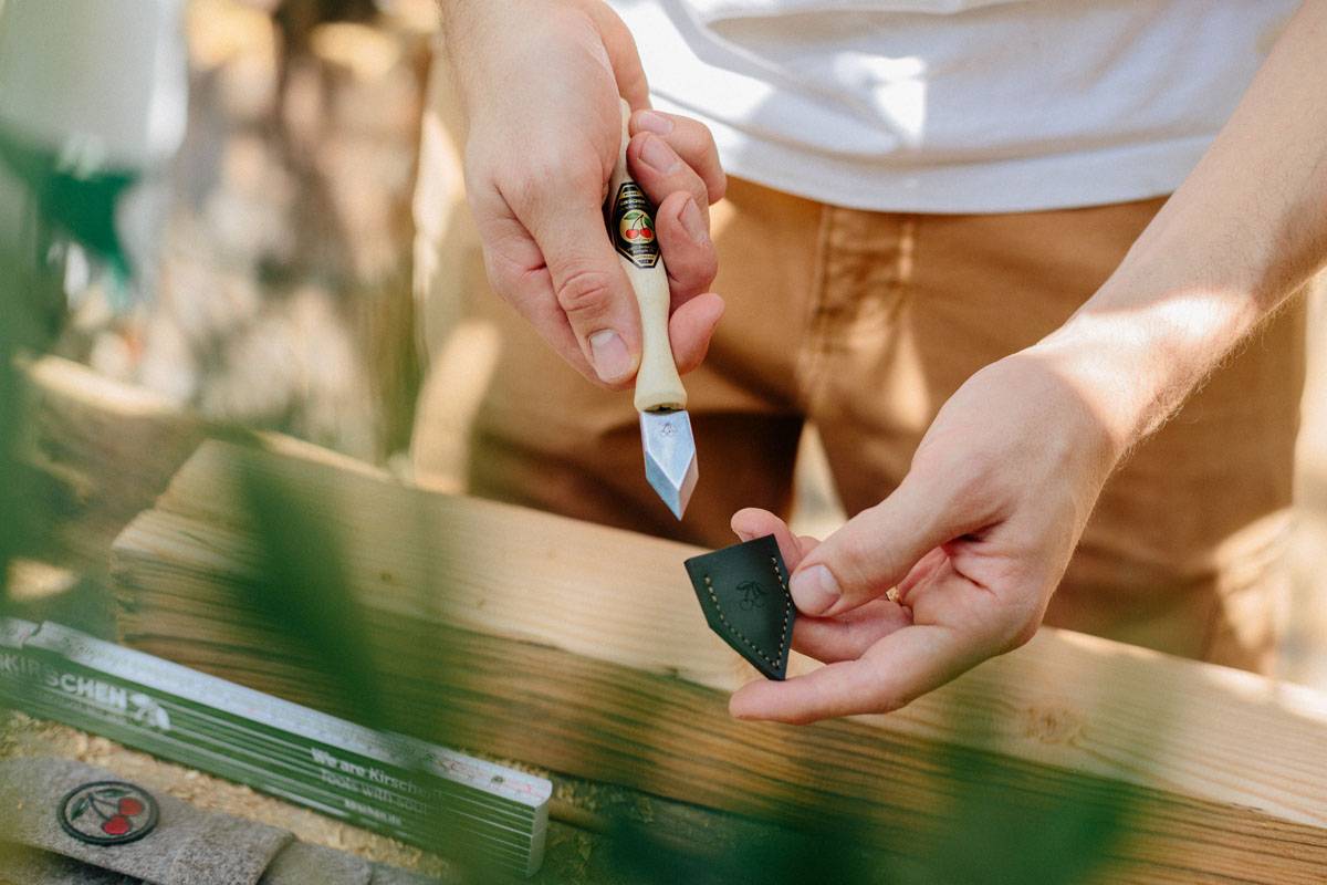 Eine Person hält ein kleines Schnitzelmesser gegen ein Stück Holz, anscheinend bereit zum Schnitzen, mit unscharfem Außenbereich im Hintergrund.