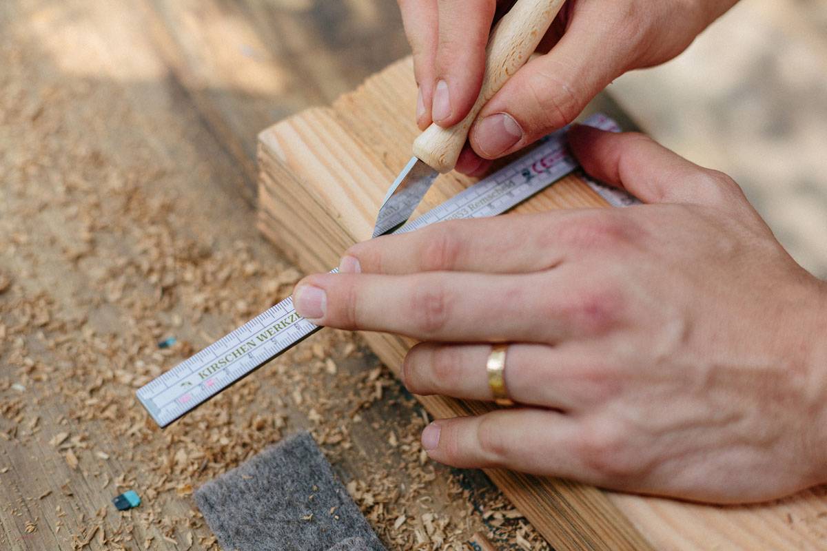 Eine Person benutzt ein Universalmesser, um Holz zu schneiden, und nutzt dazu ein Lineal für präzise Messungen. Holzspäne sind auf dem Tisch sichtbar.