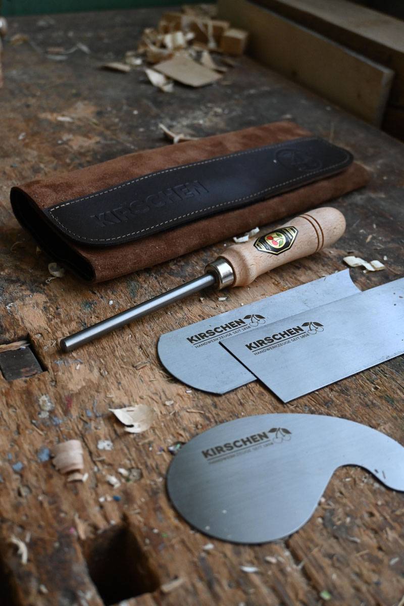 Holzbearbeitungswerkzeuge einschließlich Meißel und einer Schärfstange auf einer Werkbank aus Holz, mit einer Werkzeugrolle aus braunem Leder mit der Aufschrift „Kirschen